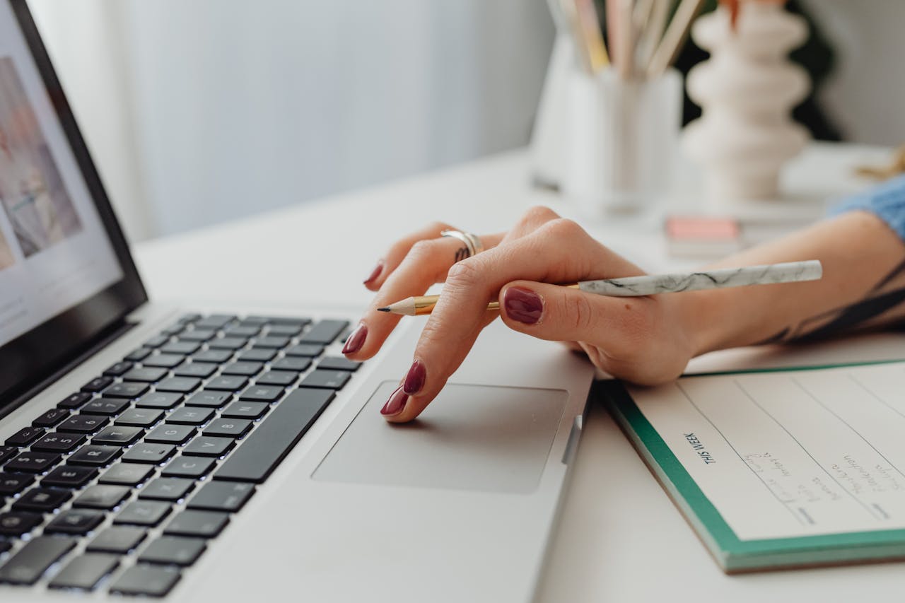 A close-up shot of a woman's hand on a laptop touchpad, holding a pencil, beside a notebook.